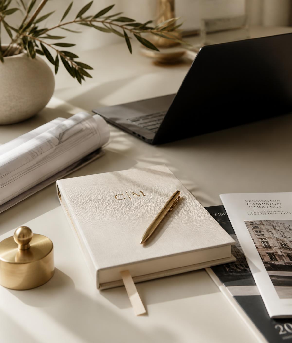 Ivory desk with black laptop, linen notebook, gold pen, campaign materials and olive branches in soft window light